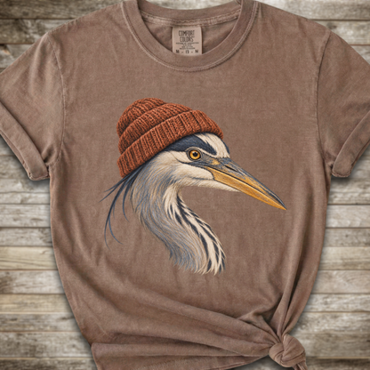 A brown short-sleeve t-shirt with a graphic of a heron wearing a brown knit beanie, styled on a rustic wooden background.