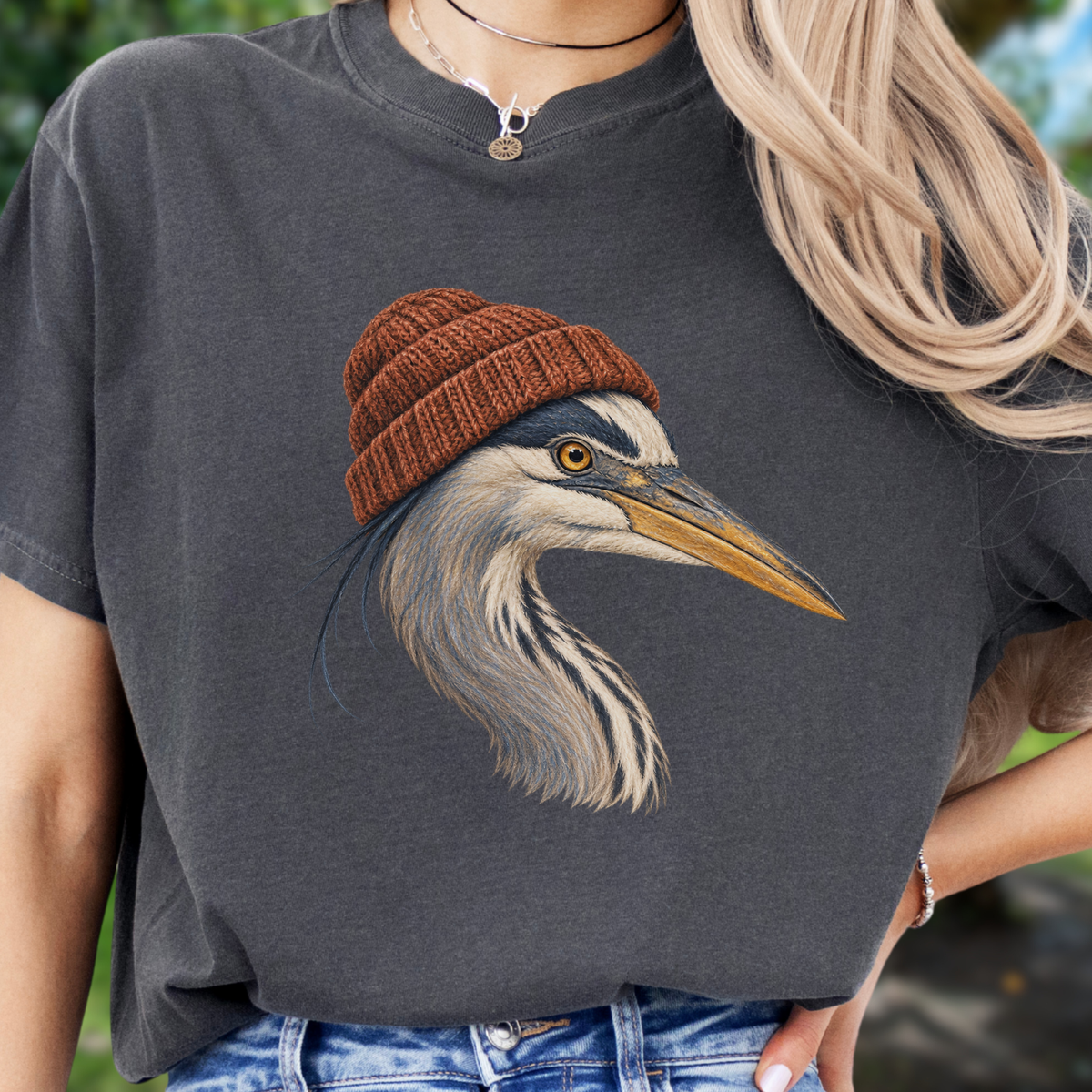A woman wearing a dark charcoal gray t-shirt with a detailed graphic of a heron wearing a brown knit beanie. The background is a soft, blurred outdoor green.