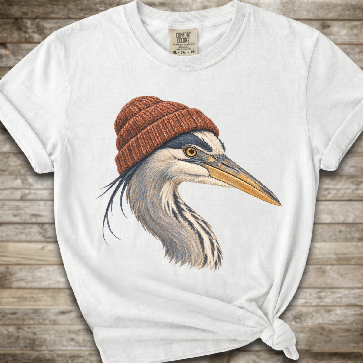 A white short-sleeve t-shirt with a graphic of a heron wearing a brown knit beanie, styled with a knot at the bottom hem against a rustic wooden background.