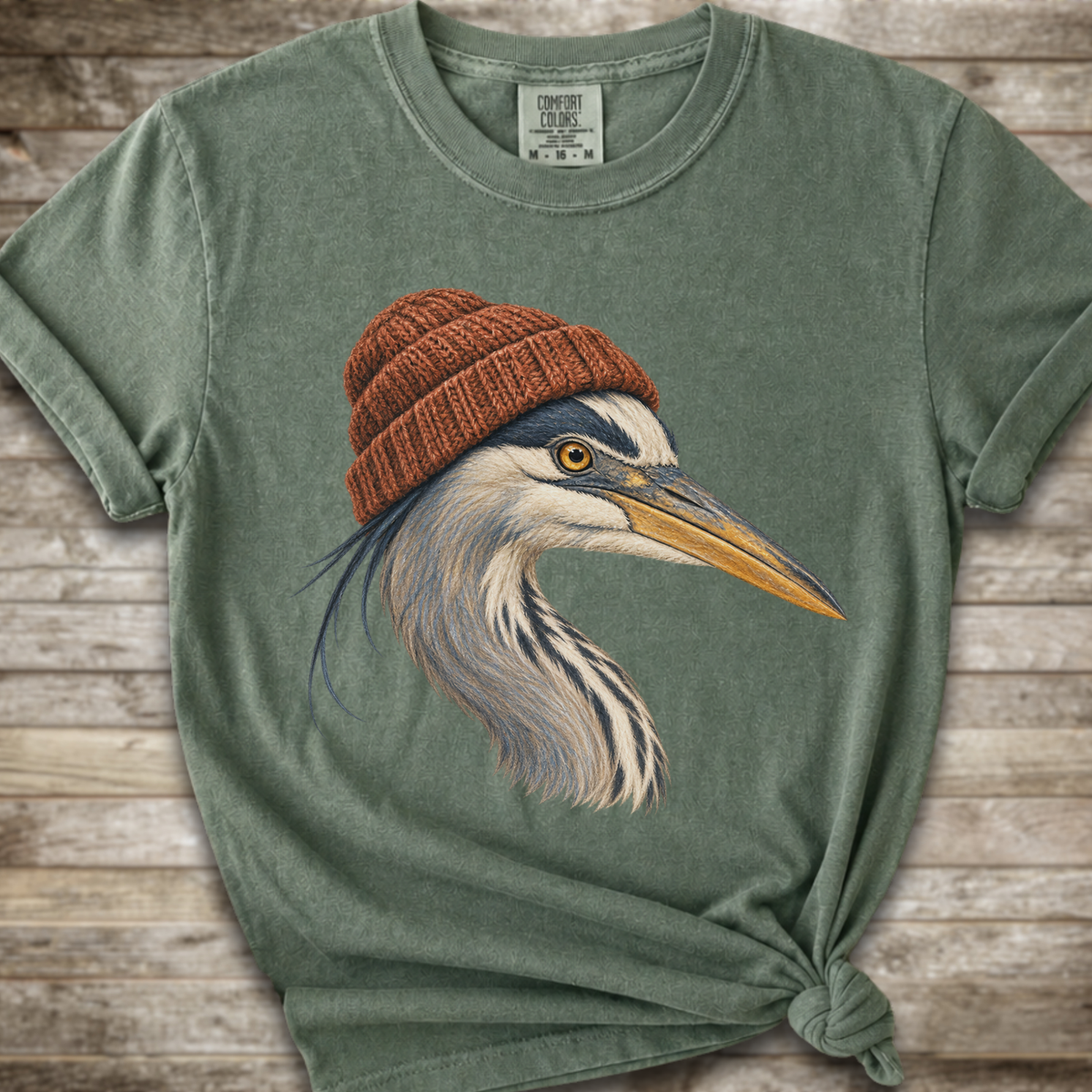 A sage green short-sleeve t-shirt with a graphic of a heron wearing a brown knit beanie, styled with a knot at the bottom hem against a rustic wood plank background.