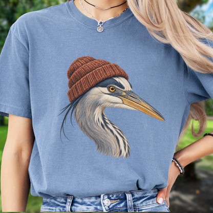 A person wearing a  blue short-sleeve t-shirt with a detailed graphic of a heron wearing a brown knit beanie. The background is a soft-focus outdoor green setting.