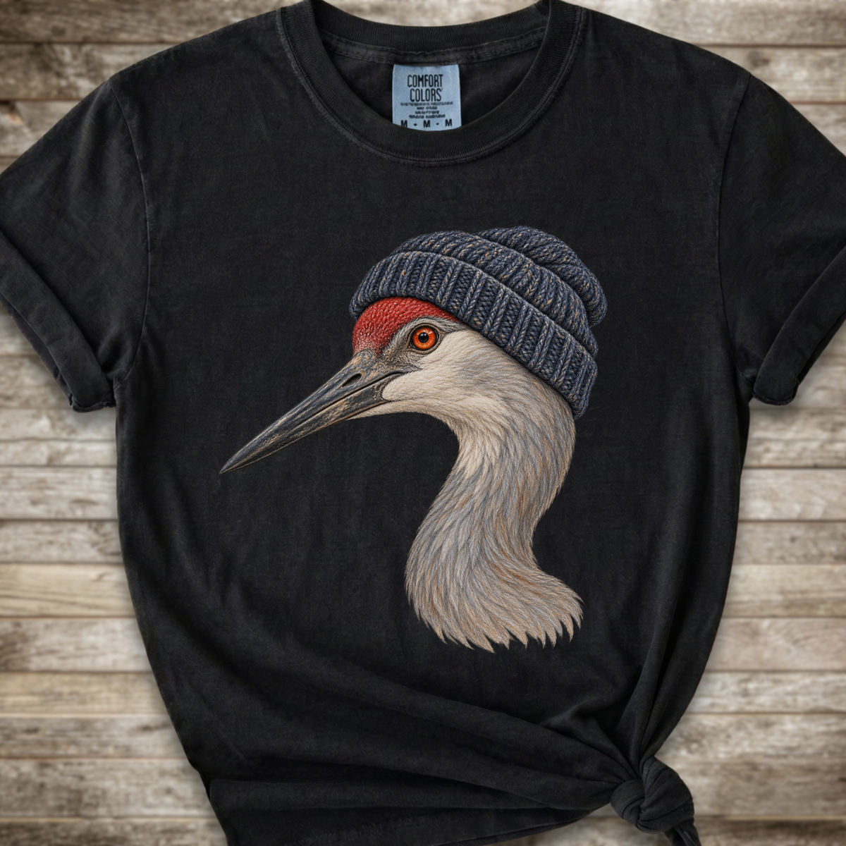A black short-sleeve t-shirt with a graphic of a crane wearing a gray knit beanie, displayed on a rustic wooden background.
