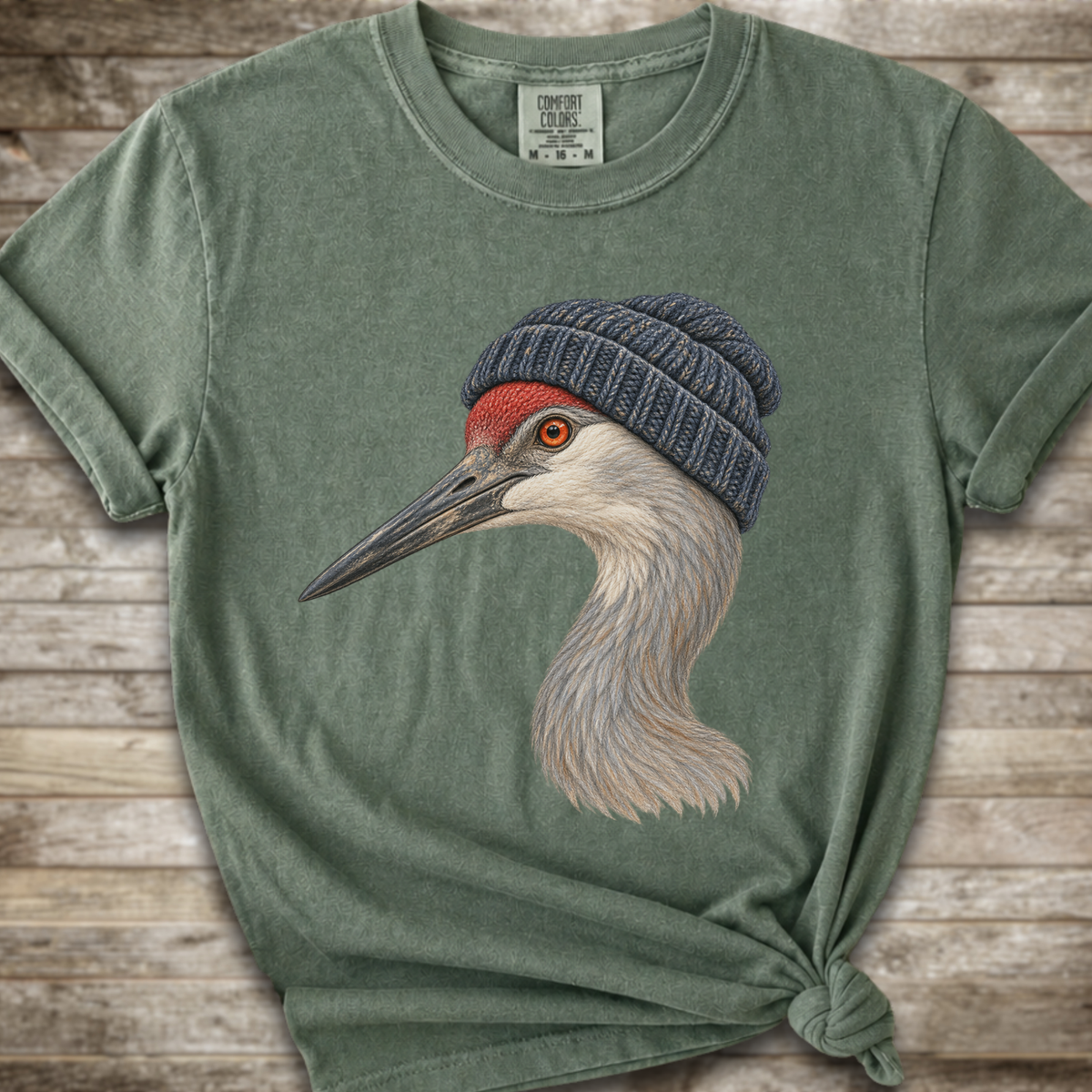 A sage green crewneck t-shirt featuring a graphic of a sandhill crane wearing a navy blue knit beanie, styled with a knot at the bottom hem against a rustic wood plank background.