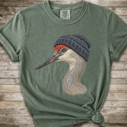 A sage green crewneck t-shirt featuring a graphic of a sandhill crane wearing a navy blue knit beanie, styled with a knot at the bottom hem against a rustic wood plank background.