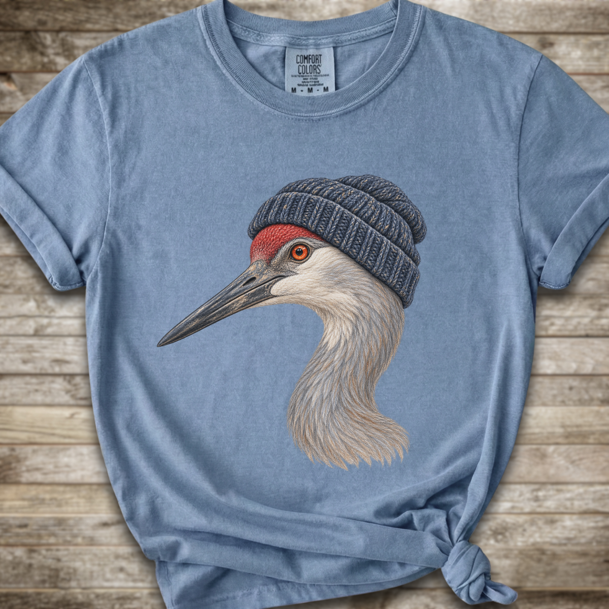 A light blue heathered t-shirt featuring a detailed graphic of a sandhill crane wearing a dark gray knit beanie, laid flat on a rustic wooden background.