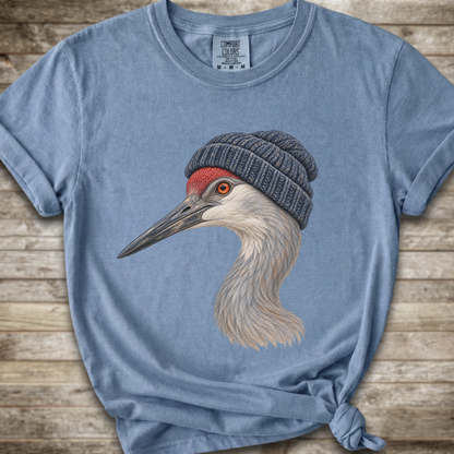 A light blue heathered t-shirt featuring a detailed graphic of a sandhill crane wearing a dark gray knit beanie, laid flat on a rustic wooden background.