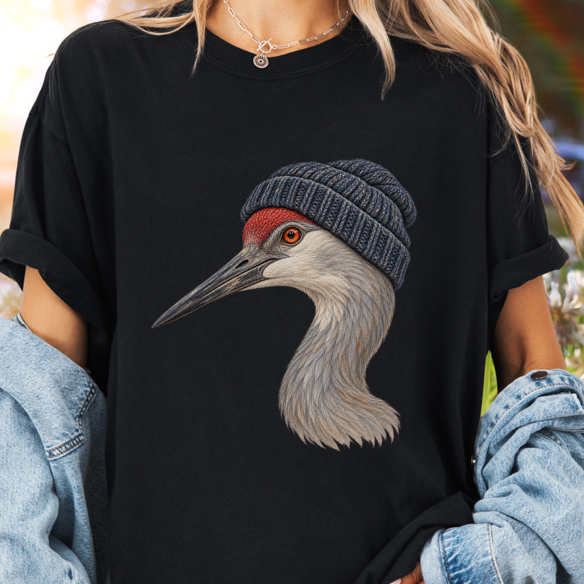 A person wearing a black oversized T-shirt with a large graphic of a crane wearing a gray knit beanie. The person has blonde hair, a silver necklace, and is wearing a light blue denim jacket draped off the shoulders.