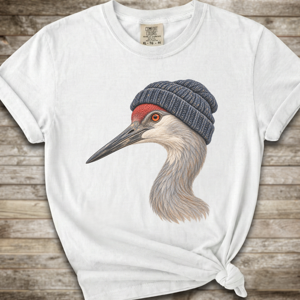 A white short-sleeve t-shirt with a graphic of a sandhill crane wearing a dark blue knit beanie, styled on a rustic wooden background with the bottom knot tied up.