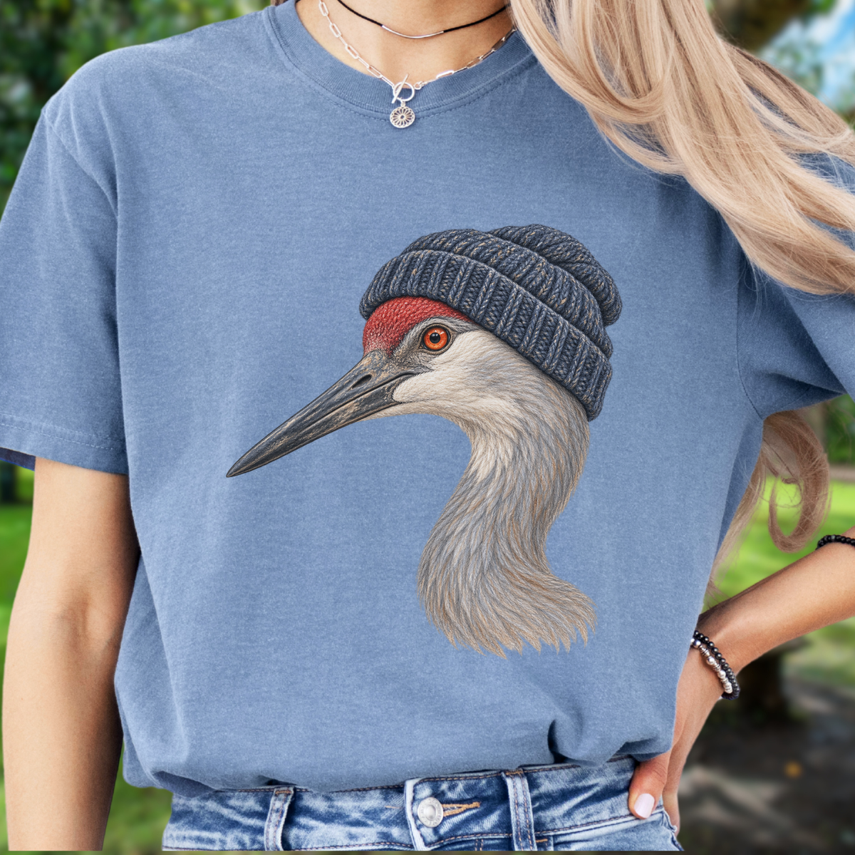 A person wearing a  blue crewneck t-shirt with a detailed graphic of a crane wearing a gray knit beanie. The background is a soft-focus outdoor green setting.