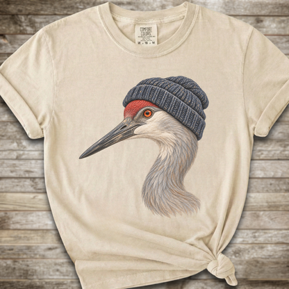 A cream-colored t-shirt with a graphic of a sandhill crane wearing a dark blue knit beanie, styled on a rustic wooden background.