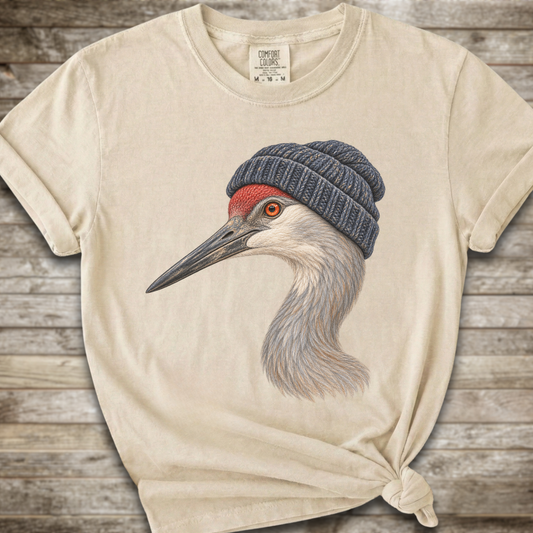 A cream-colored t-shirt with a graphic of a sandhill crane wearing a dark blue knit beanie, styled on a rustic wooden background.