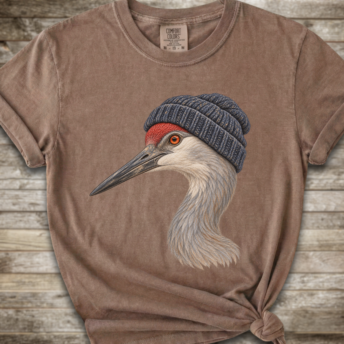 A brown short-sleeve t-shirt with a graphic of a sandhill crane wearing a blue knit beanie, displayed on a rustic wooden background.