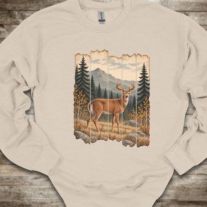 A beige crewneck sweatshirt featuring a rustic vertical wood-plank graphic print of a deer in a mountain pine forest, laid flat on a rustic wooden surface.