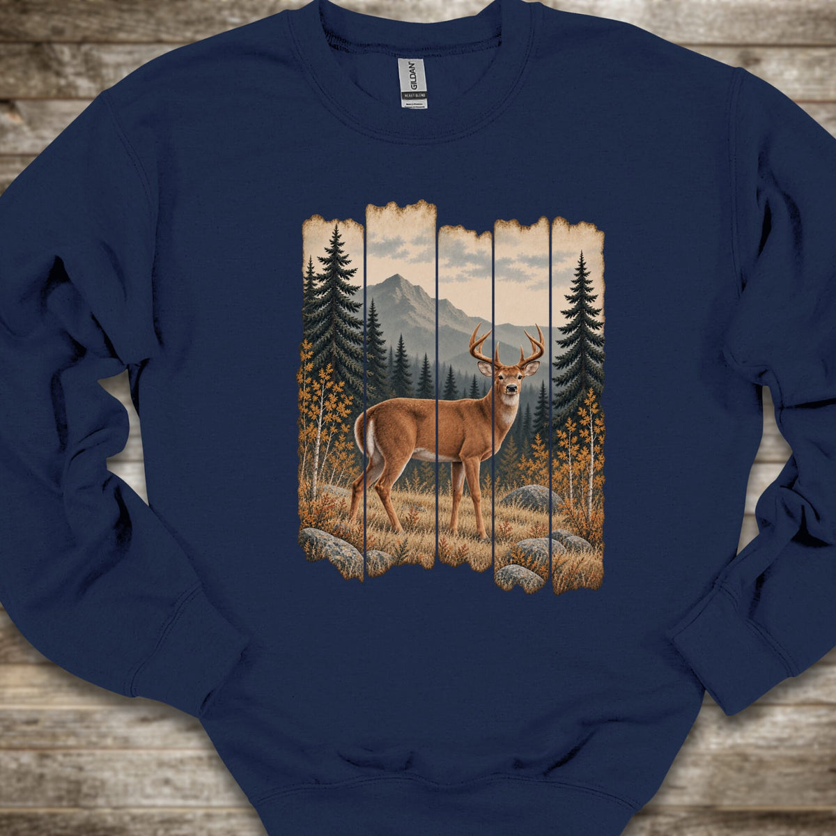 A navy blue crewneck sweatshirt featuring a rustic vertical wood-panel graphic of a deer in a forest with mountains in the background, laid flat on a rustic wooden surface.