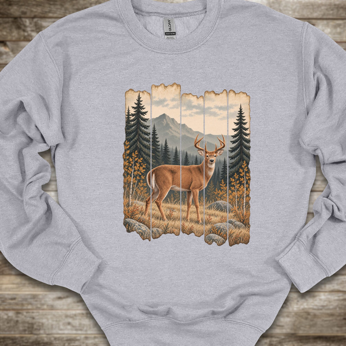 A light gray crewneck sweatshirt featuring a rustic vertical wood-plank graphic print of a deer in a mountain forest, laid flat on a rustic wooden surface.