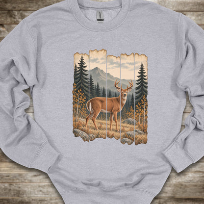 A light gray crewneck sweatshirt featuring a rustic vertical wood-plank graphic print of a deer in a mountain forest, laid flat on a rustic wooden surface.