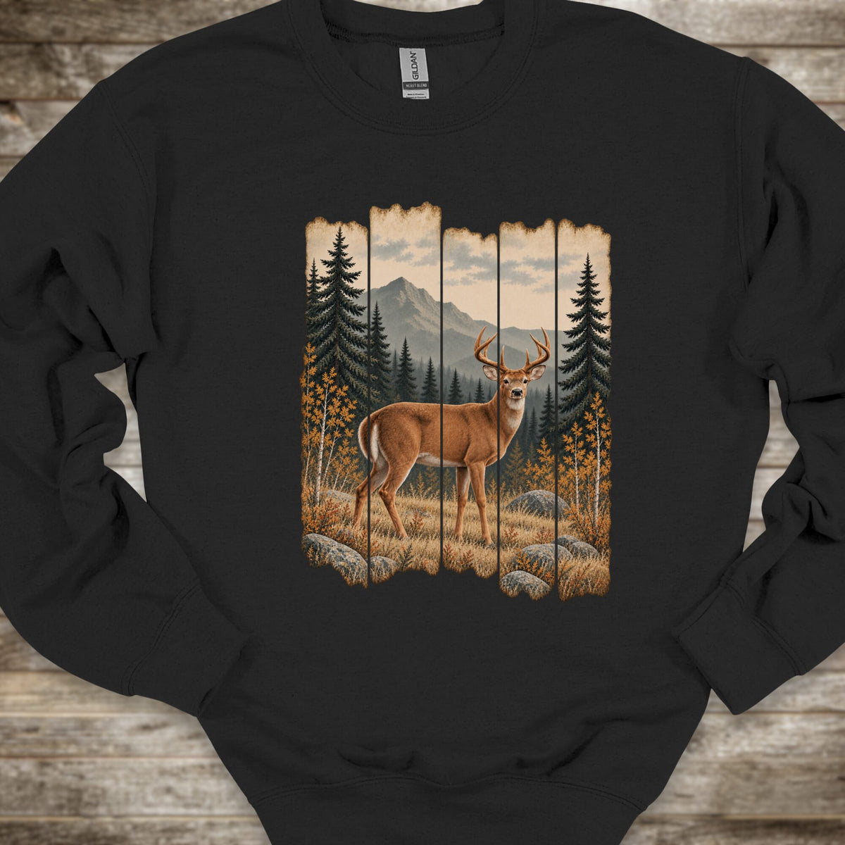 A black crewneck sweatshirt featuring a rustic vertical-panel graphic of a deer in a forest with mountains in the background, laid flat on a rustic wood plank surface.