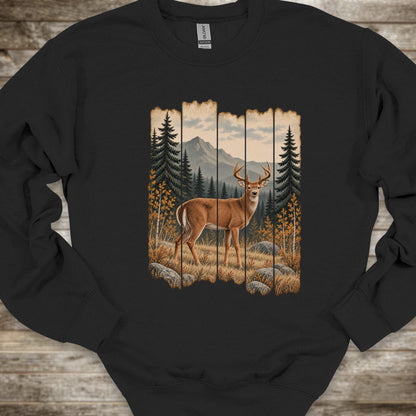 A black crewneck sweatshirt featuring a rustic vertical-panel graphic of a deer in a forest with mountains in the background, laid flat on a rustic wood plank surface.