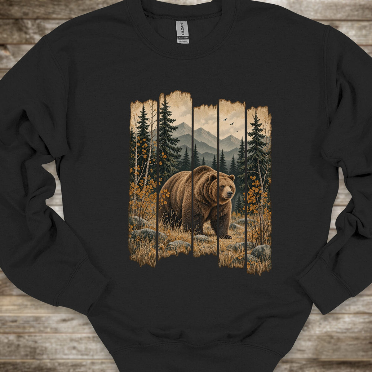A black crewneck sweatshirt featuring a large graphic print of a brown grizzly bear in a forest with mountains in the background, laid flat on a rustic wooden surface.
