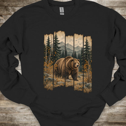 A black crewneck sweatshirt featuring a large graphic print of a brown grizzly bear in a forest with mountains in the background, laid flat on a rustic wooden surface.