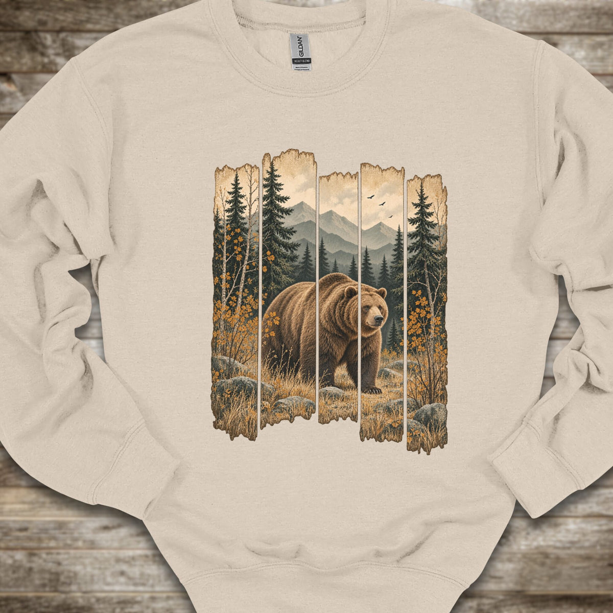 A beige crewneck sweatshirt laid flat on a rustic wooden surface, featuring a large graphic print of a brown grizzly bear walking through a forest with mountains in the background, framed by vertical wood-plank borders.