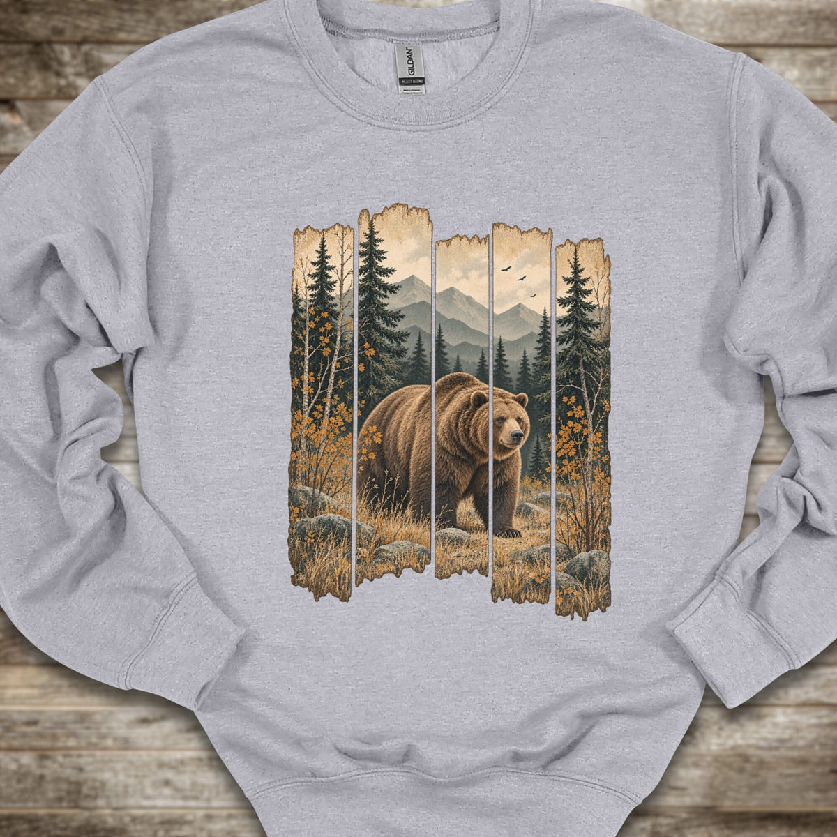 A light heather gray crewneck sweatshirt featuring a large graphic print of a brown grizzly bear in a forest scene framed by vertical wooden planks, laid flat on a rustic wooden surface.
