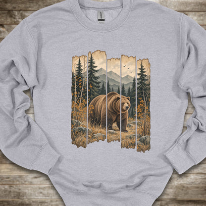 A light heather gray crewneck sweatshirt featuring a large graphic print of a brown grizzly bear in a forest scene framed by vertical wooden planks, laid flat on a rustic wooden surface.