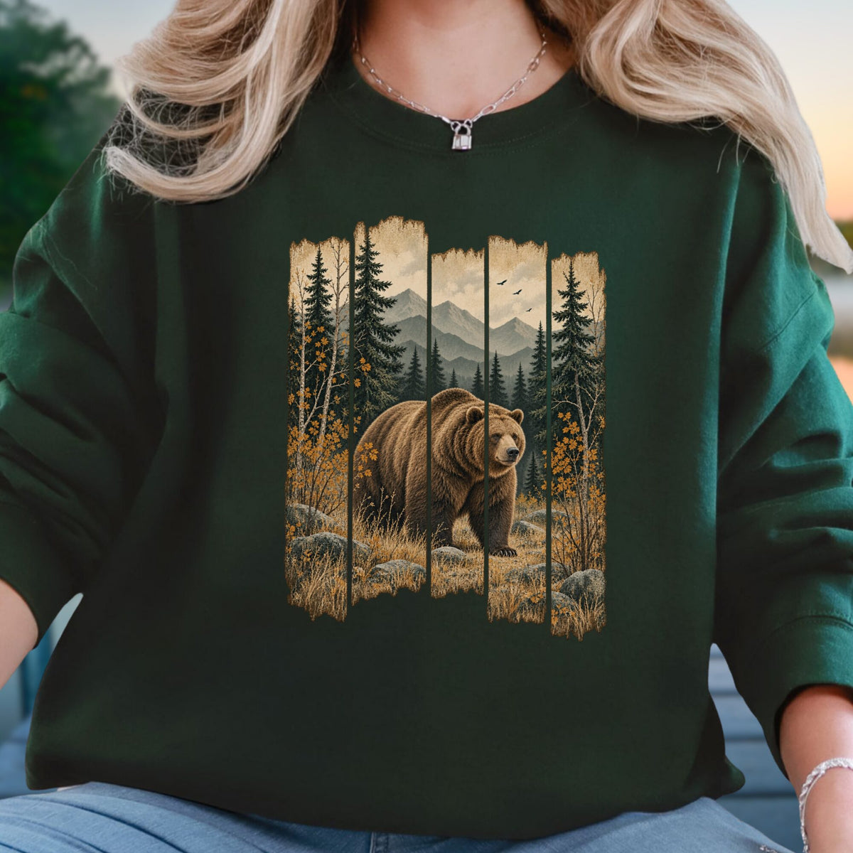 A person wearing a dark forest green crewneck sweatshirt with a large graphic print of a brown grizzly bear in a mountain pine forest. The person is also wearing blue jeans and a silver necklace.