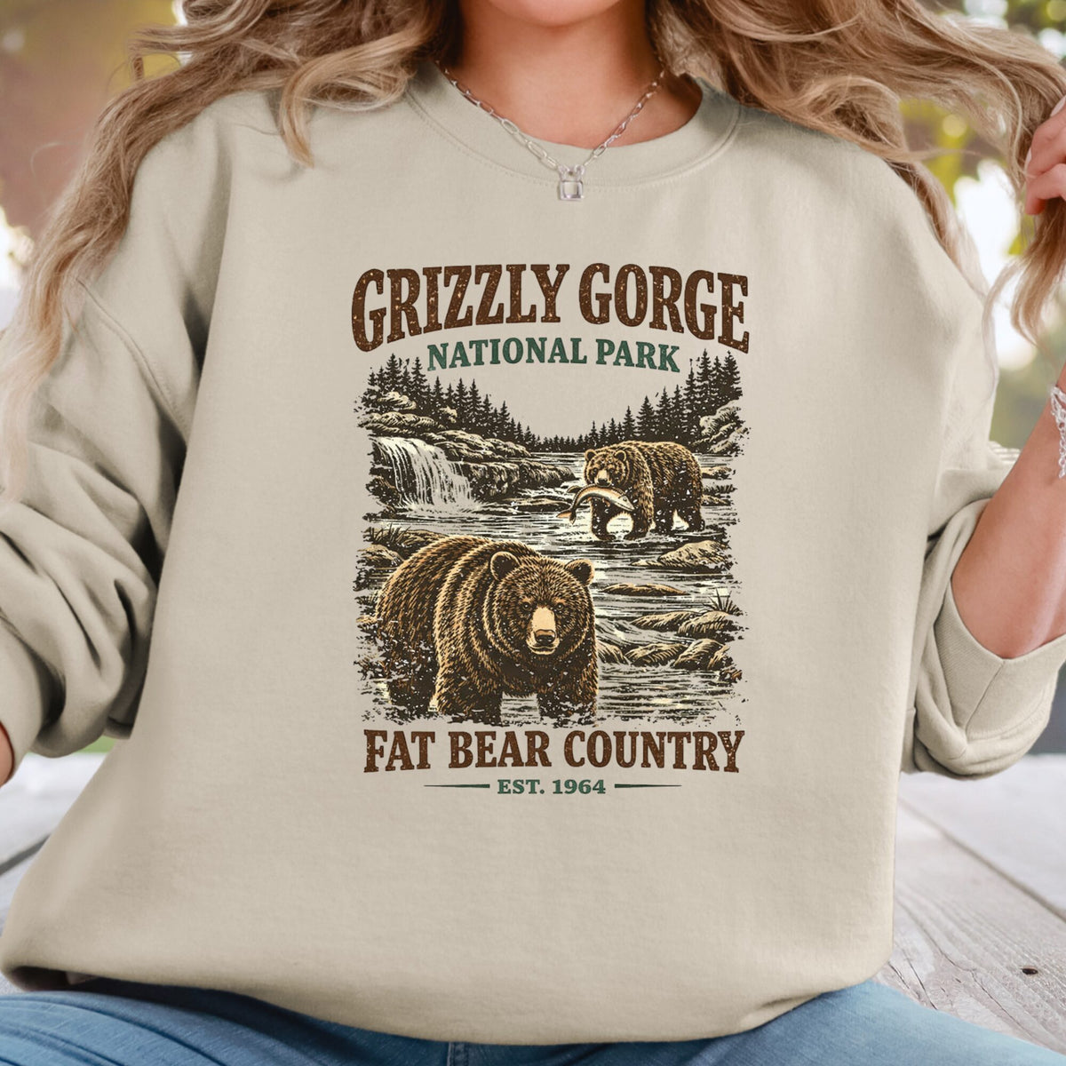 A woman wearing a beige crewneck sweatshirt with a rustic Grizzly Gorge National Park graphic featuring two bears and the text 'Fat Bear Country'.