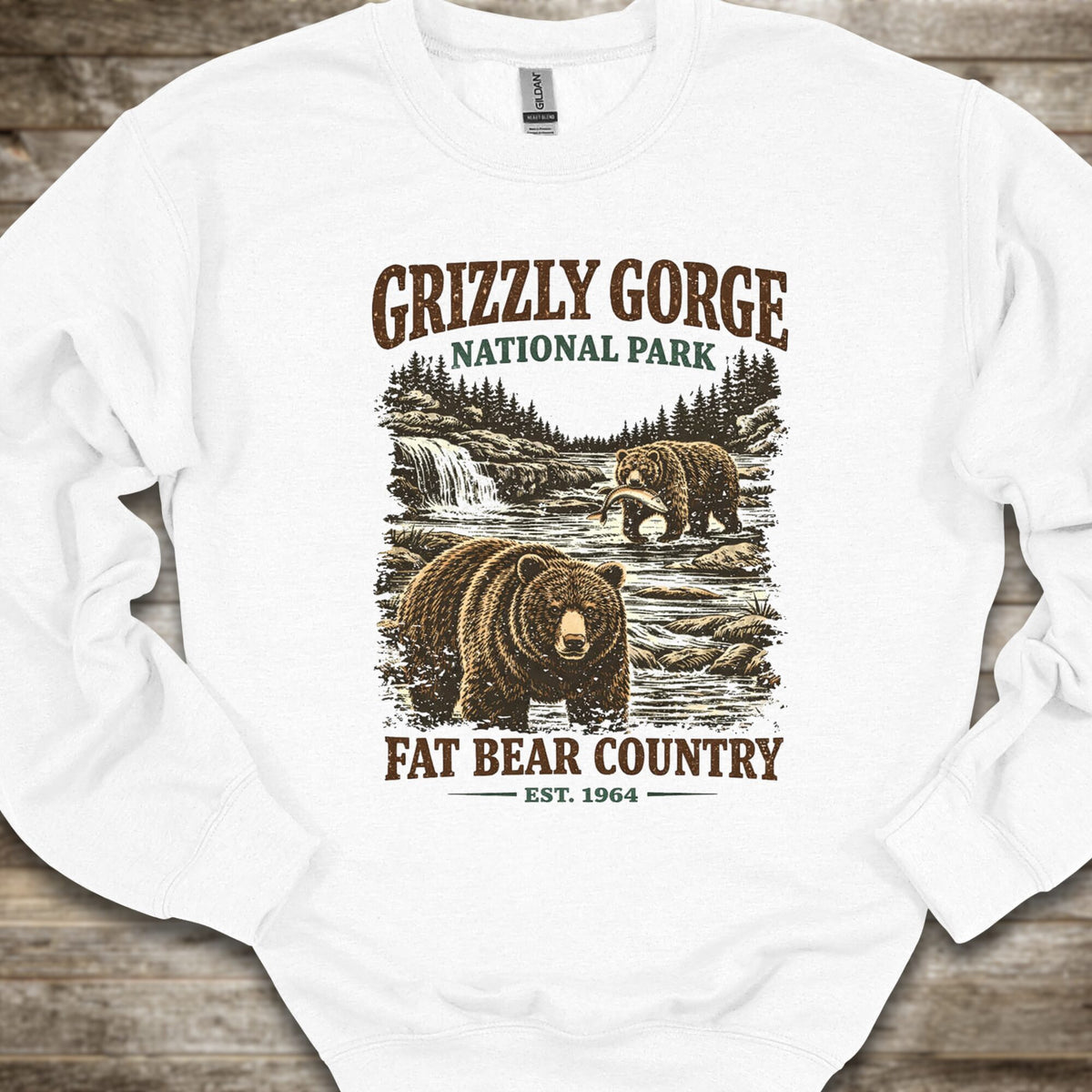 A white crewneck sweatshirt laid flat on a rustic wooden background, featuring a vintage-style graphic print of grizzly bears in Grizzly Gorge National Park with the text 'GRIZZLY GORGE NATIONAL PARK' and 'FAT BEAR COUNTRY EST. 1964'.