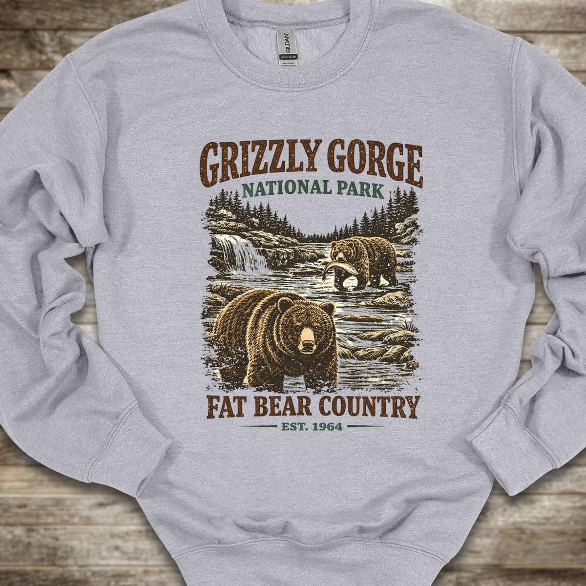 A light gray crewneck sweatshirt featuring a vintage-style graphic of grizzly bears in a national park setting with the text 'Grizzly Gorge National Park' and 'Fat Bear Country Est. 1964', laid flat on a rustic wooden background.