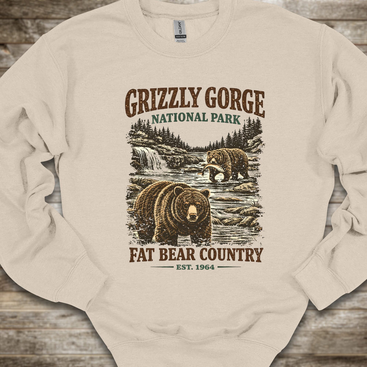 A cream-colored crewneck sweatshirt with a vintage-style Grizzly Gorge National Park graphic featuring brown bears, a waterfall, and the text 'GRIZZLY GORGE NATIONAL PARK' and 'FAT BEAR COUNTRY EST. 1964', laid flat on a rustic wooden background.