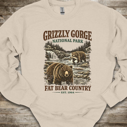 A cream-colored crewneck sweatshirt with a vintage-style Grizzly Gorge National Park graphic featuring brown bears, a waterfall, and the text 'GRIZZLY GORGE NATIONAL PARK' and 'FAT BEAR COUNTRY EST. 1964', laid flat on a rustic wooden background.