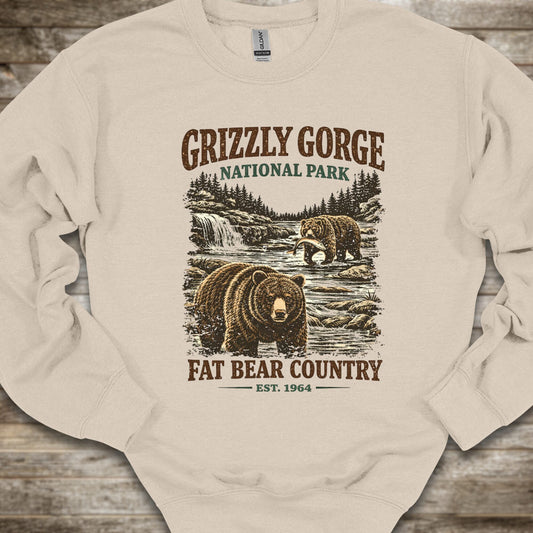 A cream-colored crewneck sweatshirt with a vintage-style Grizzly Gorge National Park graphic featuring brown bears, a waterfall, and the text 'GRIZZLY GORGE NATIONAL PARK' and 'FAT BEAR COUNTRY EST. 1964', laid flat on a rustic wooden background.