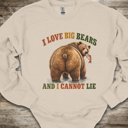 A cream-colored long-sleeve crewneck sweatshirt featuring a graphic of a brown grizzly bear holding a fish, with the text 'I LOVE BIG BEARS AND I CANNOT LIE' in a rustic orange and green font, laid flat on a rustic wooden background.