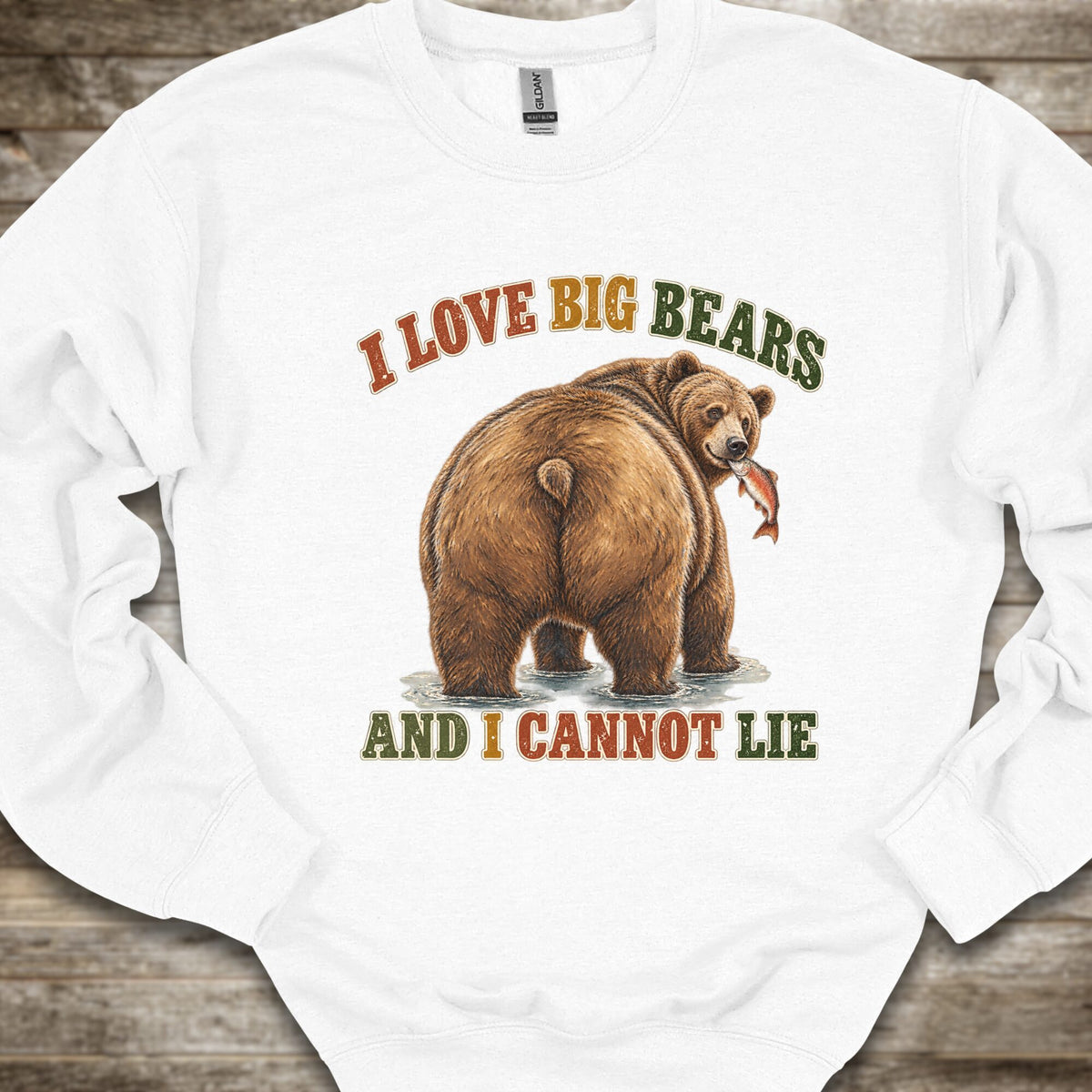 A white crewneck sweatshirt featuring a graphic of a brown grizzly bear holding a fish, with the text 'I LOVE BIG BEARS AND I CANNOT LIE' in brown and green lettering, laid flat on a rustic wooden background.