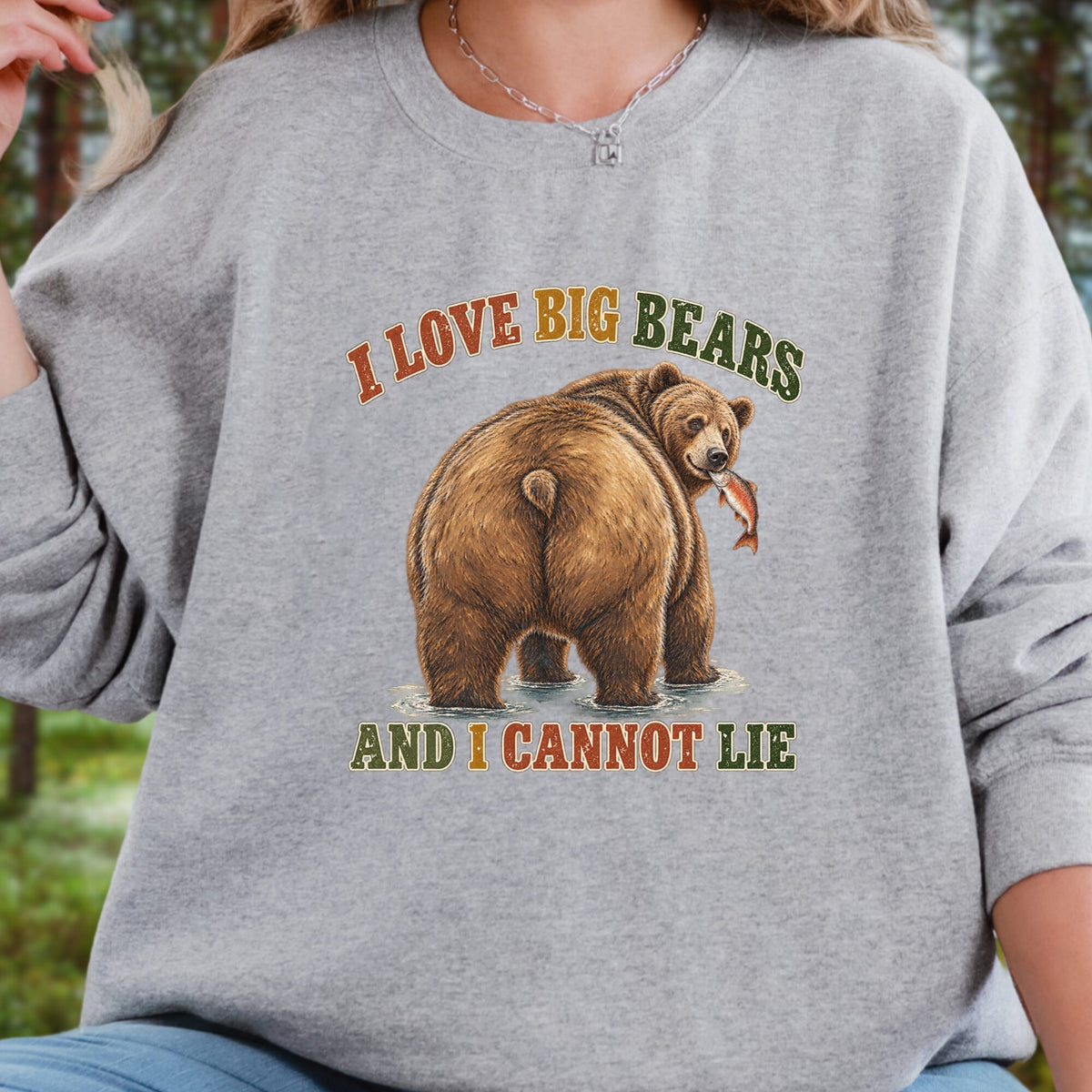 A person wearing a heather gray crewneck sweatshirt with a graphic of a brown bear holding a fish and the text 'I LOVE BIG BEARS AND I CANNOT LIE' in orange and olive green lettering, set against a blurred outdoor background.
