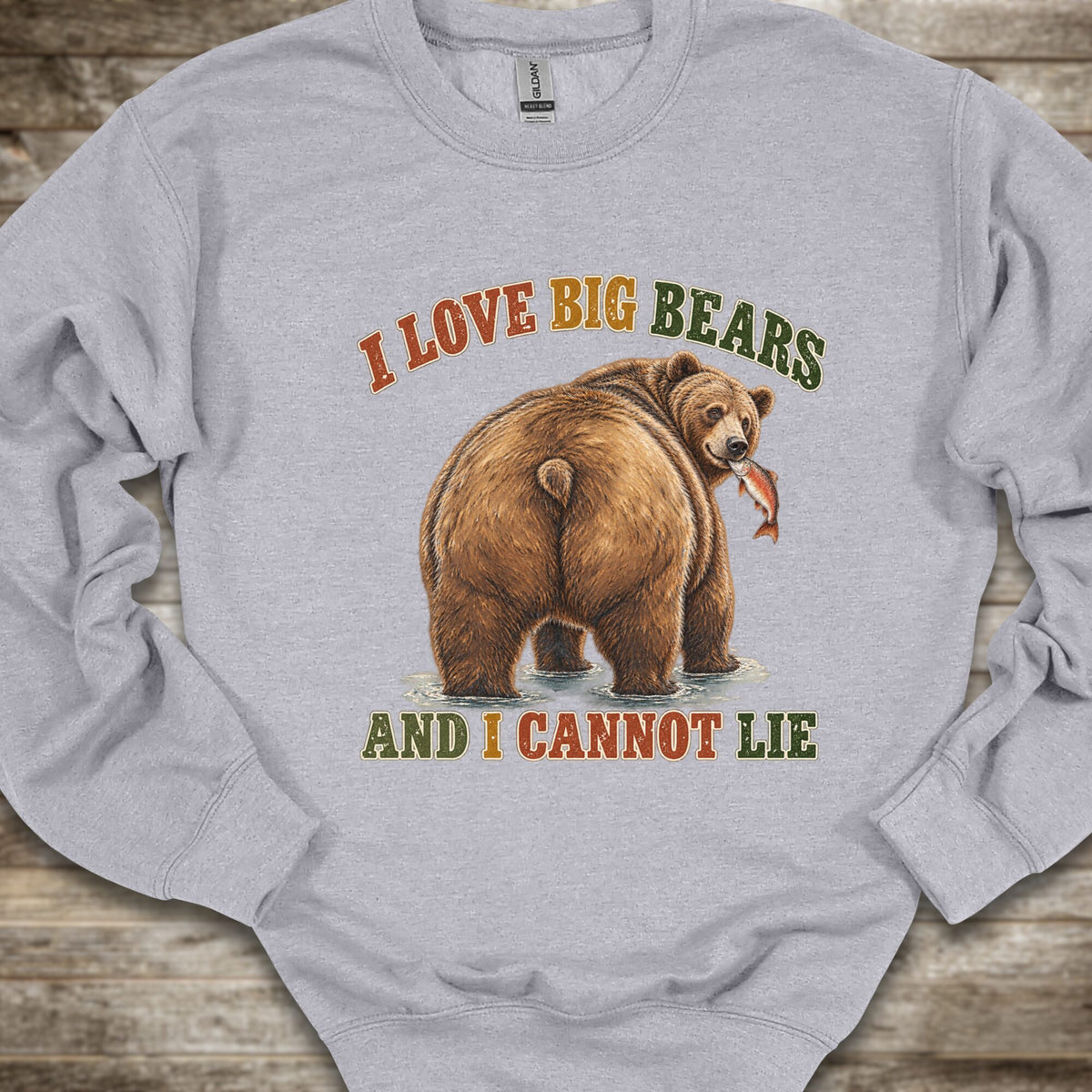 A light heather gray crewneck sweatshirt featuring a graphic of a brown grizzly bear holding a fish, with the text 'I LOVE BIG BEARS AND I CANNOT LIE' in orange and green lettering, laid flat on a rustic wooden background.