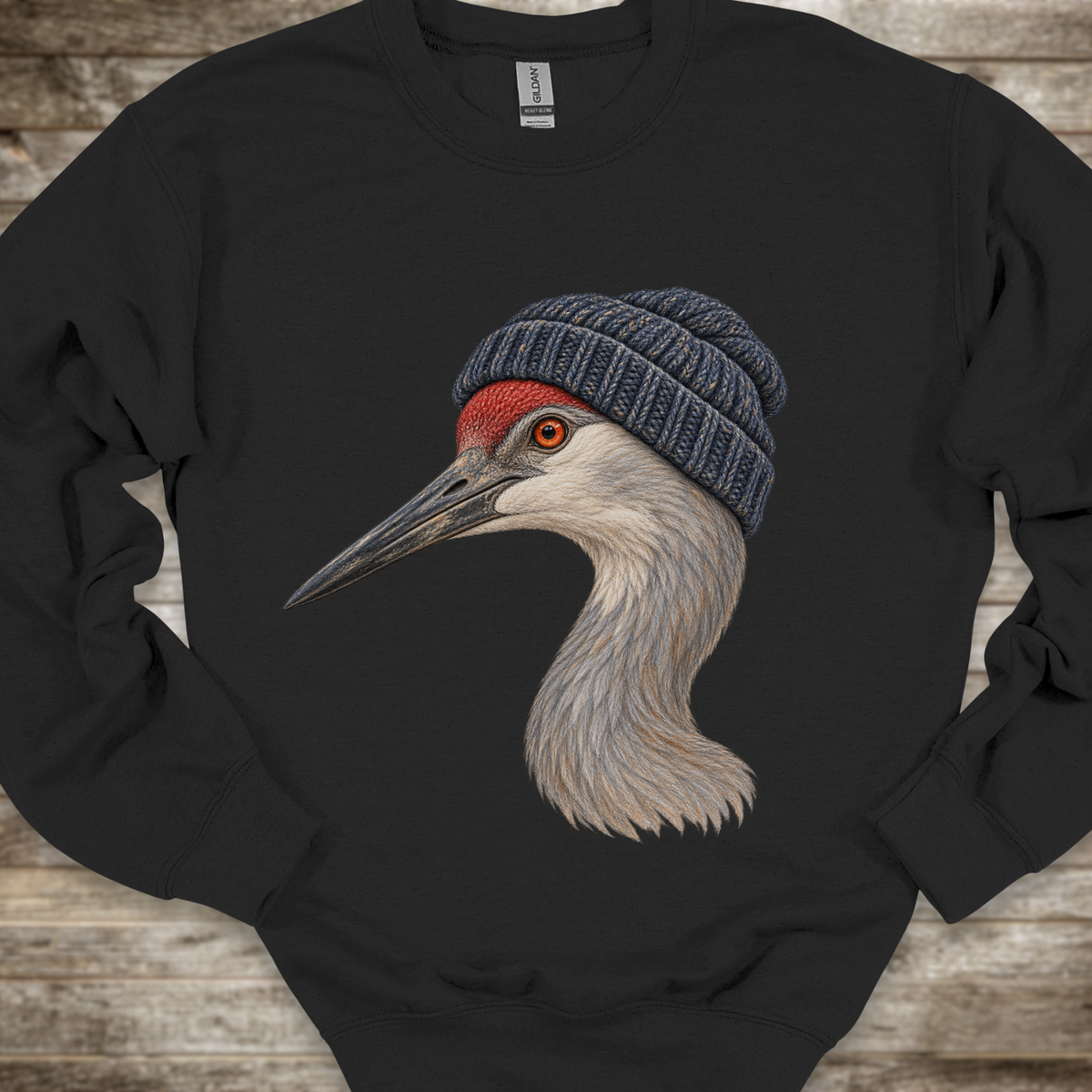 A black crewneck sweatshirt featuring a detailed graphic of a sandhill crane wearing a blue knit beanie, laid flat on a rustic wooden background.
