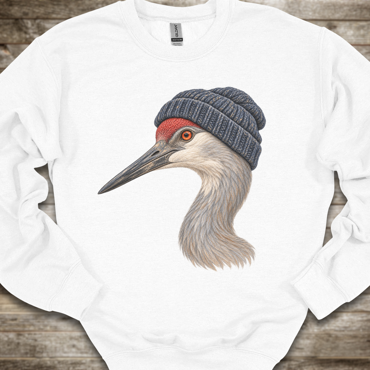 A white crewneck sweatshirt laid flat on a rustic wooden background, featuring a detailed graphic of a sandhill crane wearing a dark gray knit beanie.
