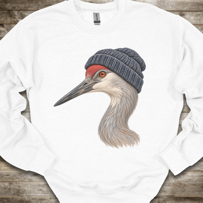 A white crewneck sweatshirt laid flat on a rustic wooden background, featuring a detailed graphic of a sandhill crane wearing a dark gray knit beanie.