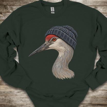 A dark forest green crewneck sweatshirt featuring a detailed graphic of a sandhill crane wearing a navy blue knit beanie, laid flat on a rustic wooden background.