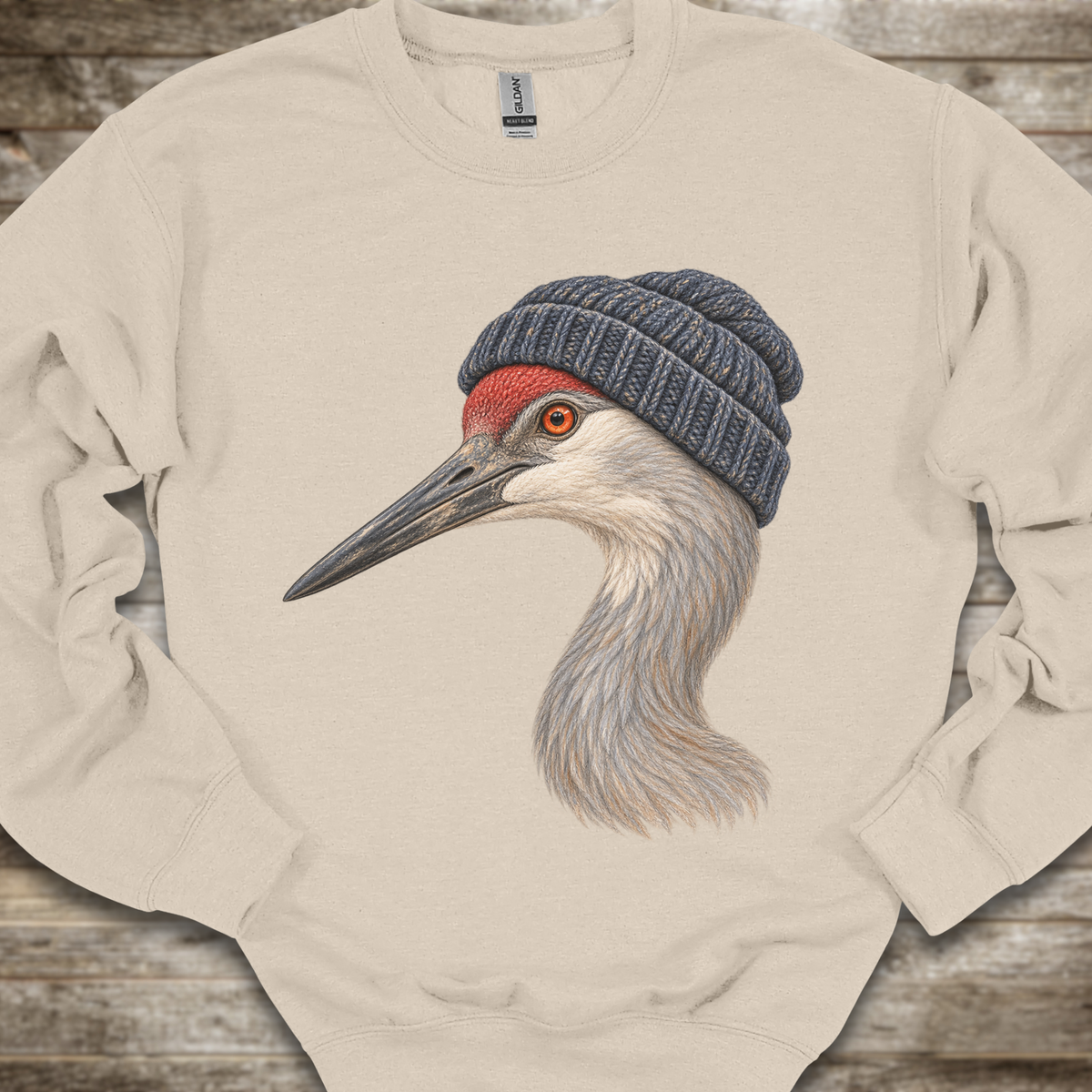 A cream-colored crewneck sweatshirt featuring a detailed graphic of a sandhill crane wearing a dark blue knit beanie, laid flat on a rustic wooden background.