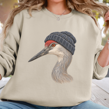 A woman wearing a beige crewneck sweatshirt with a graphic print of a sandhill crane wearing a dark blue knit beanie. The background is a soft, blurred outdoor setting.