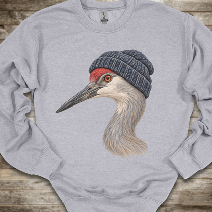 A light heather gray crewneck sweatshirt featuring a detailed graphic of a sandhill crane wearing a dark blue-gray knit beanie, laid flat on a rustic wooden surface.