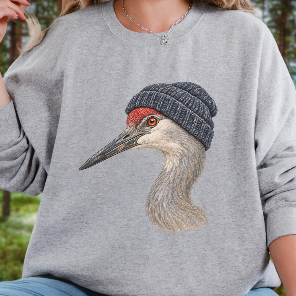A person wearing a heather gray crewneck sweatshirt with a large graphic of a crane wearing a dark knit beanie on the front. The background is a softly blurred outdoor forest setting.