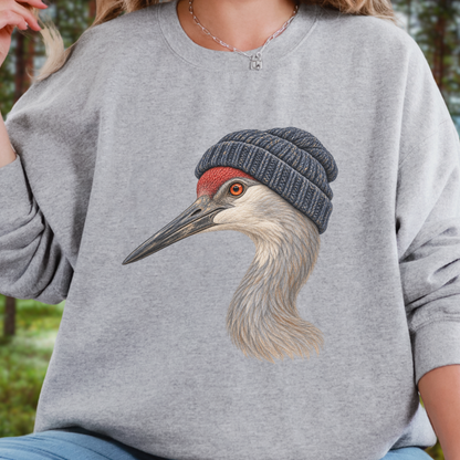 A person wearing a heather gray crewneck sweatshirt with a large graphic of a crane wearing a dark knit beanie on the front. The background is a softly blurred outdoor forest setting.