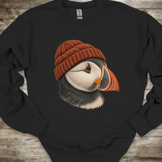 A black crewneck sweatshirt featuring a detailed graphic of a puffin wearing a brown knit beanie, laid flat on a rustic wooden background.