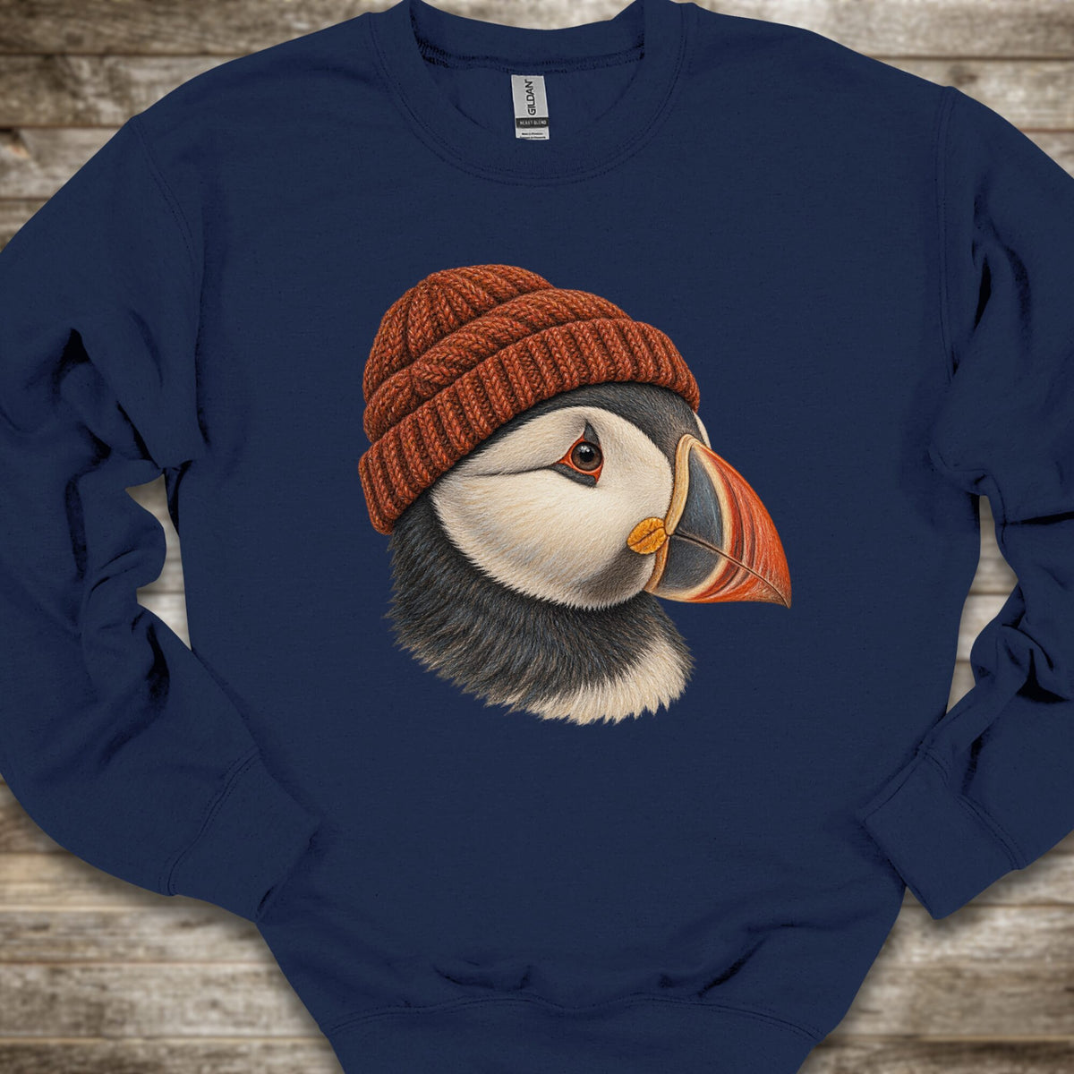 A navy blue crewneck sweatshirt featuring a detailed graphic of a puffin wearing a rust-orange knit beanie, laid flat on a rustic wooden background.