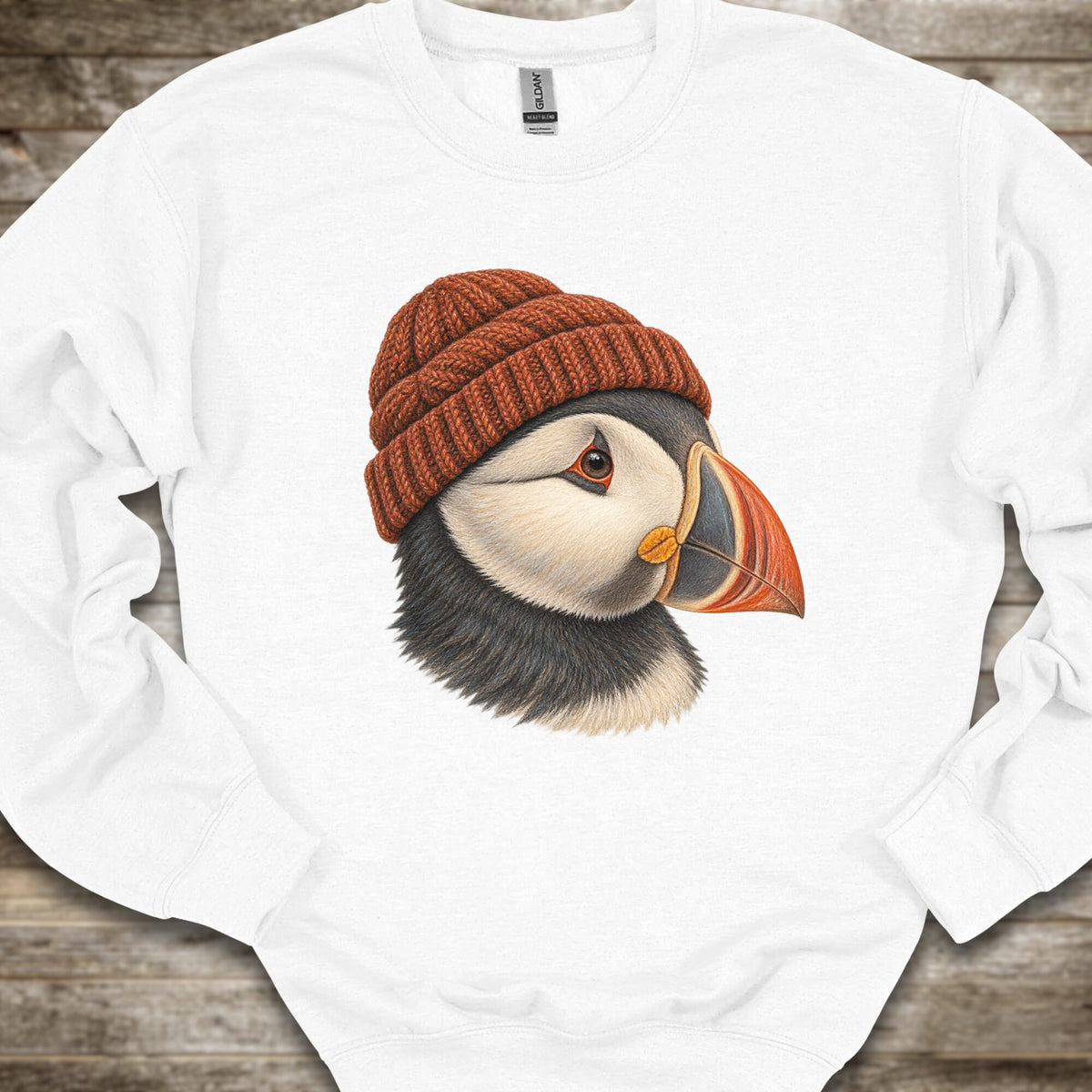 A white crewneck sweatshirt featuring a detailed graphic of a puffin wearing a brown knit beanie, laid flat on a rustic wooden background.